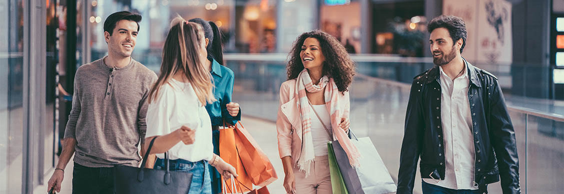 A group of boys and girls shopping together inside a bustling shopping center. They're browsing stores, carrying bags, and chatting