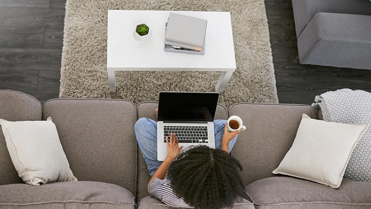 High angle shot of a young woman using a laptop and having coffee on the sofa at home