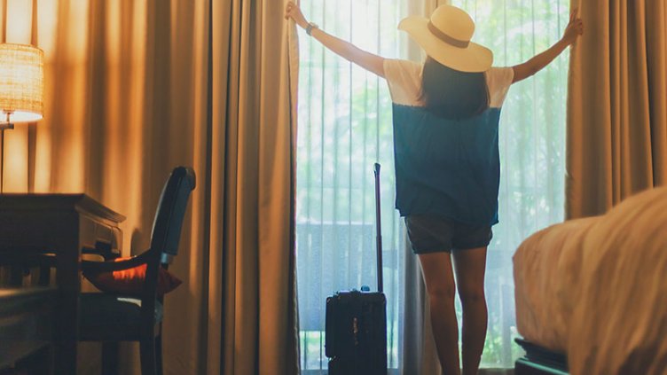 A lady looking outside while opening the curtains of a window from hotel room