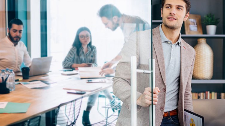 Man walking into room holding papers while people work at desks behind him